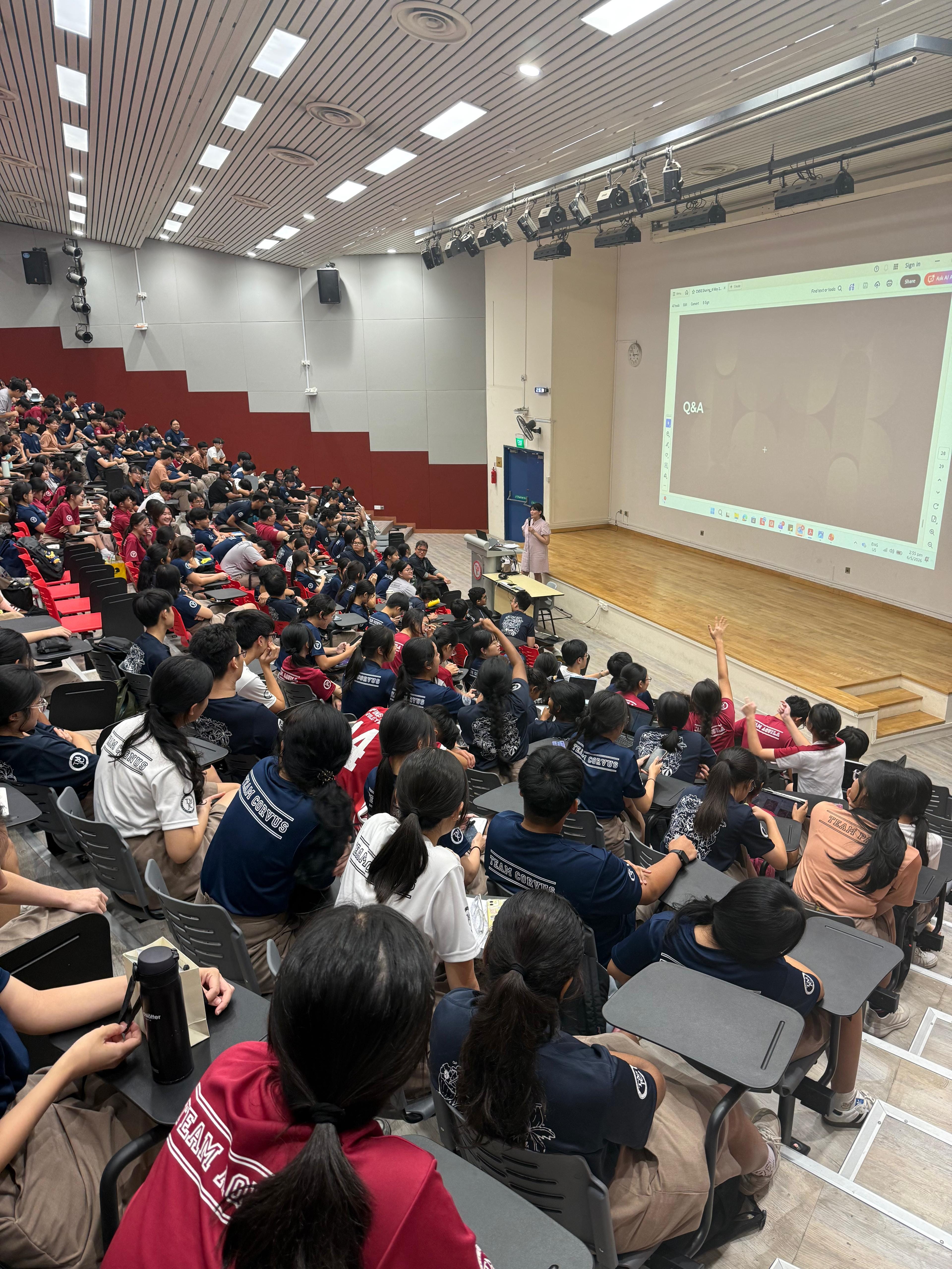 A large lecture theatre filled with students watching a presenter at the front, with a Q&A slide projected on screen and a student raising their hand.